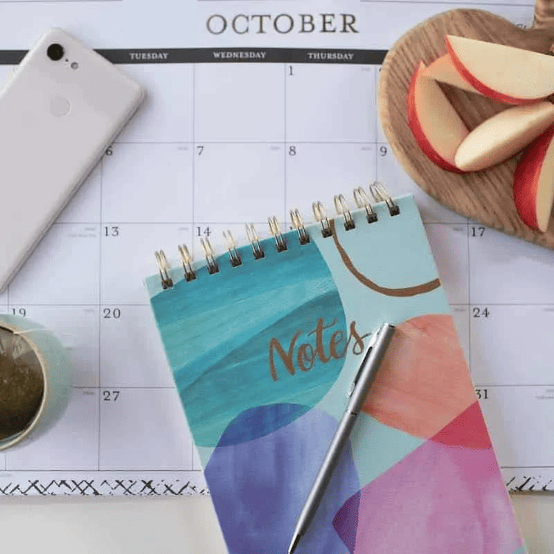 A monthly calendar with a notebook and bowl of apple slices on top.